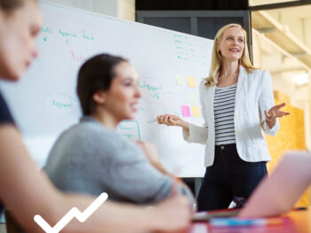 Frau in Blazer steht an einem Whiteboard und spricht in einem Meeting