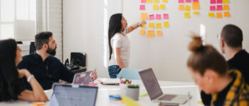 Frau mit langen Haaren steht im Meeting an einer Wand mit Post-its