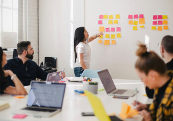 Frau mit langen Haaren steht im Meeting an einer Wand mit Post-its