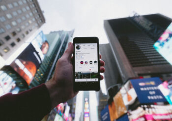 Hand hält Smartphone in den Himmel, im Hintergrund stehen Hochhäuser