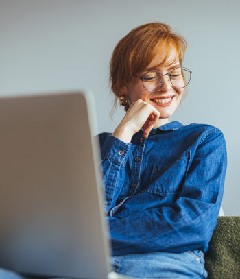 Eine Person mit roten Haaren und Brille lächelt, während sie auf einen Laptop schaut und sich auf den Start ihrer neuen Website freut. Sie trägt ein blaues Jeanshemd und sitzt mit einer Hand am Kinn vor einer schlichten, hellen Wand.