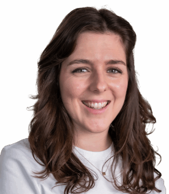 Eine Frau mit langen, gewellten braunen Haaren lächelt in die Kamera und verkörpert Selbstvertrauen und Kreativität - Qualitäten, die im Performance Marketing unerlässlich sind. Sie trägt ein weißes Oberteil und eine zarte Halskette vor einem hellgrauen Hintergrund.