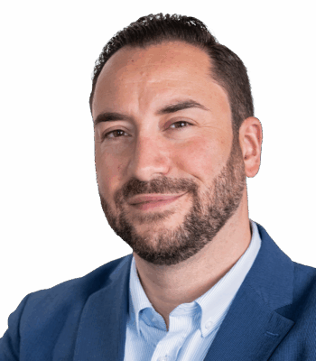 Ein Performance Manager mit kurzen dunklen Haaren und gestutztem Bart, der einen blauen Blazer über einem hellblauen Hemd trägt, blickt leicht lächelnd in die Kamera vor einem schlichten hellen Hintergrund.