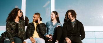 Vier junge Erwachsene sitzen draußen zusammen, lächeln und unterhalten sich. In lässigen Jacken und Jeans strahlen sie die Wärme einer starken Gemeindeverwaltung aus. Im Hintergrund fällt Sonnenlicht durch ein großes Fenster.