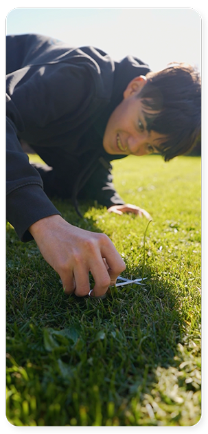 Ein junger Mensch liegt an einem sonnigen Tag lächelnd im Gras, während er den Boden genau betrachtet oder berührt und die Natur in einer vom Barrierefreiheitsgesetz geprägten Umgebung genießt.