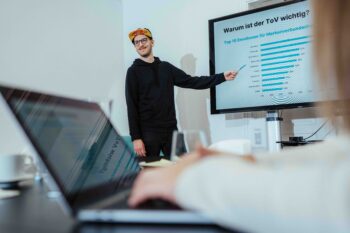 Ein Mann mit schwarzem Kapuzenpullover und Kappe hält einen Vortrag zum Thema Community Management und zeigt auf einen Bildschirm mit einem deutschen Balkendiagramm, während jemand im Vordergrund auf einem Laptop tippt.