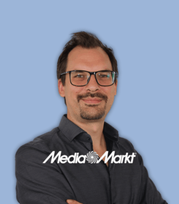 Ein Mann mit Brille, dunklem Haar und dunklem Hemd steht mit verschränkten Armen da und lächelt in die Kamera. Im unteren Bildbereich sind das MediaMarkt-Logo und „Community Management“ eingeblendet. Der Hintergrund ist hellblau.