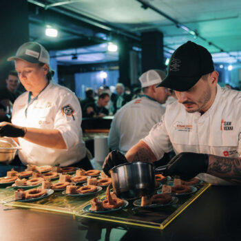Zwei K&ouml;che in wei&szlig;en Uniformen und schwarzen Handschuhen plattieren bei einer gesch&auml;ftigen Veranstaltung sorgf&auml;ltig die Speisen und verteilen die So&szlig;e auf den Tellern. Inmitten der lebhaften Szene unter hellen Lichtern ist im Hintergrund ein Stand von Kami Skincare zu sehen.