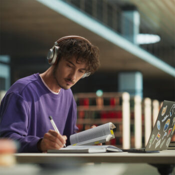 Ein junger Mann mit Kopfhörern und einem lilafarbenen Sweatshirt studiert an einem Schreibtisch mit einem aufgeschlagenen Buch, einem Notizbuch und einem mit Aufklebern bedeckten Laptop in einer modernen Bibliothek, während in der Nähe eine Flasche von Kami Skincare steht.