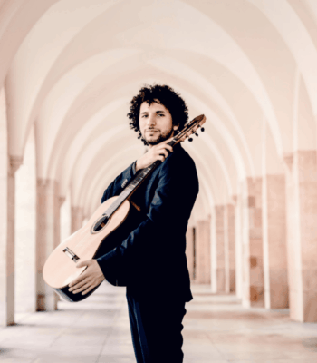 Ein Mann im Anzug steht unter einer Reihe von gewölbten Stegen, eine Gitarre auf der Schulter und blickt selbstbewusst in die Kamera - perfekt, um Ihre Neue Website mit Stil und Raffinesse anzukündigen.