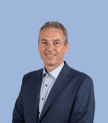 Ein Mann in einem marineblauen Anzug und einem hellblauen Hemd lächelt in die Kamera. Er hat kurzes graues Haar und steht vor einem schlichten hellblauen Hintergrund, perfekt für die Verwendung auf einer professionellen Website.