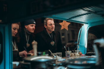 Drei Personen mit herzförmigen Halsketten stehen an einem blauen Imbisswagen und schauen aufmerksam zu, was gerade zubereitet wird. Die Szene ist warm beleuchtet, auf der Theke stehen Gläser und Utensilien, darüber hängt ein Holzstern.