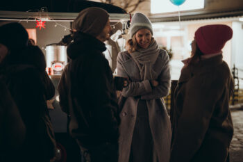 Eine Gruppe von winterlich gekleideten Menschen steht nachts draußen, unterhält sich und lächelt in der Nähe eines mit Lichterketten geschmückten Imbisswagens. Eine Frau in einem grauen Mantel und mit Mütze steht in der Mitte und unterhält sich.