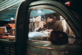 Ein Mann und eine Frau in Winterkleidung bestellen nachts in einem Imbisswagen Essen, gesehen durch das Fenster des Wagens. Die Szene ist warm beleuchtet und schafft eine gemütliche Atmosphäre.