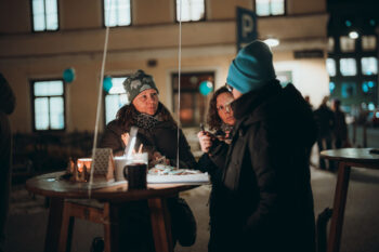 Drei Personen in Winterkleidung stehen nachts um einen Holztisch im Freien, unterhalten sich und essen. Das warme Licht von Kerzen und Gebäuden schafft eine gemütliche Atmosphäre. Der Hintergrund zeigt eine sanft beleuchtete Straße und ein Parkplatzschild.