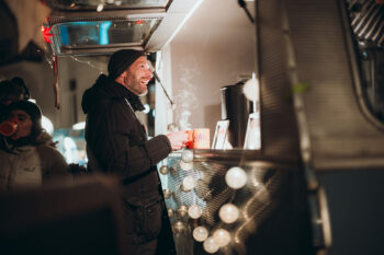Ein Mann in Wintermantel und Mütze lächelt, während er einen dampfenden Becher an einem mit Lichterketten geschmückten Imbisswagen hält. Eine andere Person im Hintergrund trinkt aus einer roten Tasse. Die Szene wirkt warm und festlich.