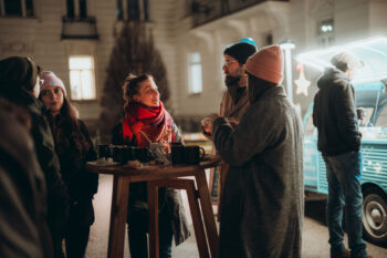 Eine Gruppe von Menschen in Winterkleidung steht um einen hohen Holztisch mit Getränken und Snacks und unterhält sich nachts im Freien in der Nähe eines Food Trucks mit hellen Lichtern.