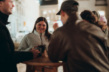 Eine Frau in einer weißen Jacke lächelt, während sie sich mit drei Personen an einem Holztisch im Freien unterhält, die Tassen halten. Der Hintergrund ist sanft mit verschwommenen Lichtern der Stadt beleuchtet.