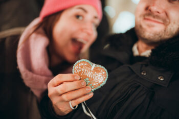 Eine lächelnde Person hält ein herzförmiges Lebkuchengebäck mit der Aufschrift Spatzl in der Hand, während eine andere Person mit rosa Mütze und Schal im Hintergrund lächelt. Beide wirken glücklich und sind für das kalte Wetter gekleidet.