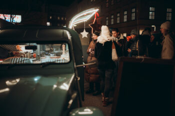 Eine Gruppe von Menschen in Winterkleidung versammelt sich bei einem nächtlichen Imbisswagen, der von festlichen sternförmigen Lichtern beleuchtet wird, während im Hintergrund Gebäude zu sehen sind.