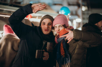 Zwei Personen in Winterjacken und Mützen posieren spielerisch bei einer Veranstaltung im Freien, lächeln und halten Tassen. Helle Lichter und verschwommene Menschen im Hintergrund suggerieren eine festliche, lebhafte Atmosphäre.