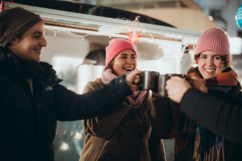 Drei Personen in Winterkleidung und Mützen lächeln und stoßen mit Tassen an, die von festlichen Lichterketten umgeben sind.
