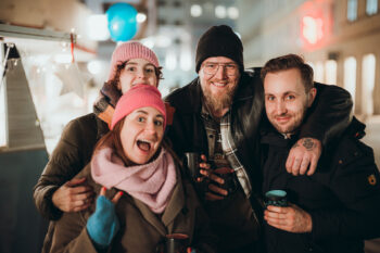 Vier Freunde in Winterkleidung lächeln und halten Tassen, während sie dicht beieinander in einer nächtlichen Stadt stehen, mit festlichen Lichtern und verschwommenen Gebäuden im Hintergrund.
