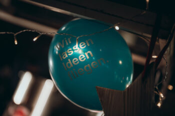 Ein blauer Luftballon mit der Aufschrift Wir lassen Ideen fliegen. ist von einer Lichterkette und einem Holzstern umgeben und schafft eine festliche Atmosphäre im Haus.