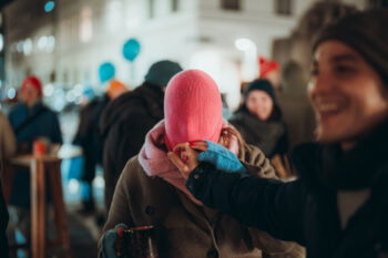 Eine Person, die eine rosafarbene Strickmütze über das ganze Gesicht gezogen hat, steht nachts im Freien, umgeben von Menschen in Winterkleidung. Eine andere Person in der Nähe lacht und berührt die Mütze. Die Szene wirkt lebendig und verspielt.