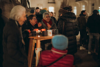 Eine Gruppe von Menschen in warmer Kleidung versammelt sich nachts im Freien um einen hohen Holztisch mit Bechern SPiNNpunsch und Kerzen und unterhält sich auf einem scheinbar festlichen Ereignis oder Markt.