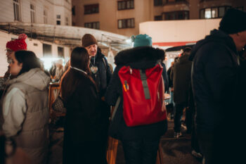 Winterlich gekleidete Menschen versammeln sich auf einem Abendmarkt im Freien. Im Hintergrund sind Stände mit Lichtern zu sehen. Eine Person mit einem leuchtend roten Rucksack steht in der Menge, und Gebäude umgeben die Szene.