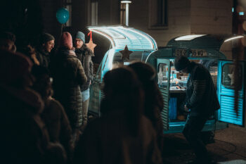 Eine Gruppe von Menschen in Winterkleidung steht nachts um einen blauen SPiNNpunsch-Imbisswagen, der von warmen Lichtern beleuchtet wird. Eine Person serviert Essen aus dem offenen Wagen, während andere in der Nähe warten und sich unterhalten. Ein Luftballon ist an dem Wagen befestigt.