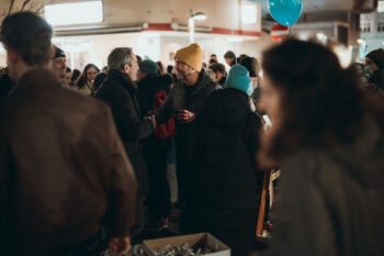 Eine Gruppe von Menschen sitzt nachts im Freien, viele in Winterkleidung, und genießt SPiNNpunsch. Zwei Männer in der Mitte schütteln sich die Hände und lächeln. Ein blauer Luftballon schwebt über der Menge und Lichter erhellen die festliche Szene.