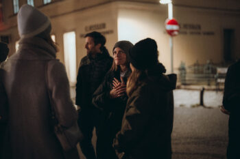 Eine Gruppe winterlich gekleideter Menschen steht nachts im Freien zusammen und unterhält sich bei SPiNNpunsch. Eine Person in der Mitte gestikuliert mit einer Hand auf ihrer Brust. Straßenlaternen und ein Gebäude leuchten sanft im Hintergrund.