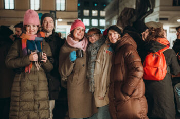 Eine Gruppe von fünf Personen in warmer Winterkleidung mit bunten Mützen und Schals lächelt und hält SPiNNpunsch-Tassen in der Hand, während sie bei einer Abendveranstaltung im Freien dicht beieinander stehen, während sich andere im Hintergrund versammeln.
