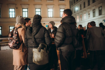 Eine Gruppe von Menschen in warmer Winterkleidung steht nachts vor einem Gebäude, plaudert und lächelt in einer gut beleuchteten städtischen Umgebung. Einige von ihnen tragen Mützen und Rucksäcke.
