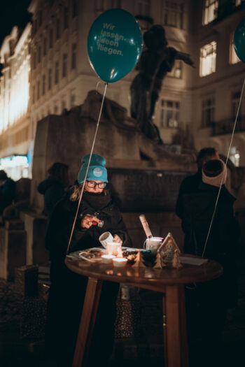 Eine Person mit blauem Hut und Mantel steht nachts an einem runden Tisch im Freien und bereitet ein Essen oder ein Getränk vor, mit Kerzen und zwei blauen Luftballons. Im Hintergrund sind eine Statue und Gebäude zu sehen.