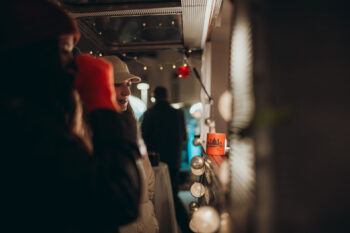 Zwei warm gekleidete Personen stehen nachts neben einem warm beleuchteten Imbisswagen oder einer Imbissbude, eine hält ein Getränk in der Hand. Lichterketten und ein rotes Ornament schaffen eine festliche, gemütliche Atmosphäre.