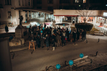Eine große Gruppe von Menschen versammelt sich nachts im Freien in der Nähe einer Statue auf einem Stadtplatz, einige halten blaue Luftballons. Tische und ein Imbisswagen sind zu sehen, die umliegenden Gebäude sind sanft beleuchtet.