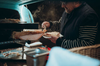 Eine Person in einem gestreiften Pullover schmilzt an einem Imbissstand mit einem Raclette-Grill Käse auf Brot, wobei Dampf vom heißen Käse aufsteigt.