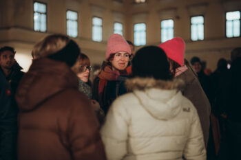 Eine Gruppe von Menschen mit Wintermänteln und Mützen versammelt sich nachts im Freien. Eine Person mit rosa Hut und Brille steht in der Mitte, lächelt und spricht mit den anderen. Im Hintergrund ist ein Gebäude mit beleuchteten Fenstern zu sehen.