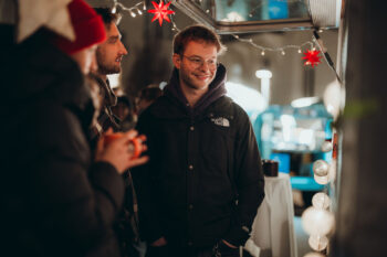 Ein Mann mit Brille und schwarzer Jacke lächelt, während er mit zwei Personen auf einem festlichen Markt im Freien steht, der mit Lichterketten und roten Sternen geschmückt ist.