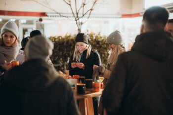 Eine Gruppe von Menschen in Winterkleidung versammelt sich um einen Holztisch mit Tassen im Freien. Zwei Frauen in der Mitte lächeln, eine schaut auf ihr Telefon. Die Atmosphäre ist locker und freundlich.