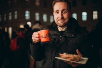 Ein Mann in einem dunklen Wintermantel hält einen orangefarbenen Becher in der einen und einen Teller mit Essen in der anderen Hand und lächelt leicht. Er steht nachts im Freien, mit verschwommenen Menschen und Gebäuden im Hintergrund.