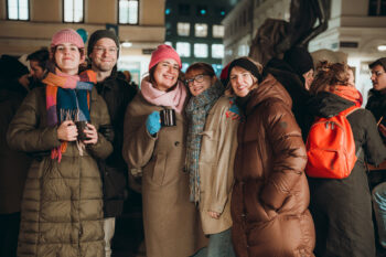 Fünf Personen in Wintermänteln, Mützen und Schals lächeln und posieren gemeinsam bei einer nächtlichen Veranstaltung im Freien. Einige halten Tassen, andere tragen Brillen. Im Hintergrund sind weitere Personen und Lichter zu sehen.