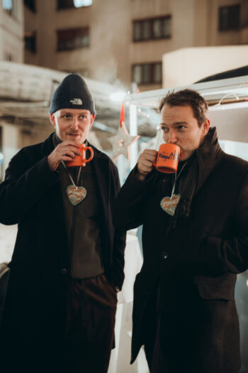 Zwei Männer in dunklen Wintermänteln und Mützen stehen im Freien, jeder hält einen leuchtend roten Becher in der Hand und nippt daraus. Sie tragen herzförmige Lebkuchenhalsketten und scheinen einen festlichen, kühlen Abend zu genießen.