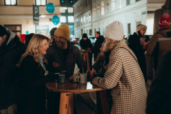 Eine Gruppe warm gekleideter Menschen steht auf einem Nachtmarkt im Freien um einen Holztisch herum, lacht und unterhält sich. Im Hintergrund sind blaue Luftballons und die Lichter der Stadt zu sehen.