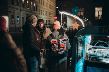 Drei Personen in warmer Kleidung stehen an einem kleinen Imbisswagen auf einer nächtlichen Straße. Eine Person trägt einen bunten Rucksack. Der Wagen ist beleuchtet und mit einem Stern und einem blauen Luftballon geschmückt. Gebäude säumen den Hintergrund.
