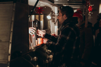 Ein Mann in einer karierten Jacke erhält ein heißes Getränk von einem Verkäufer an einem festlich geschmückten Imbisswagen, der nachts mit Lichterketten und roten Sternen verziert ist.