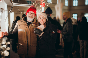Ein lächelndes älteres Paar in warmer Winterkleidung posiert zusammen bei einer festlichen Veranstaltung im Freien, die mit Lichtern geschmückt ist, wobei andere Personen im Hintergrund verschwimmen.
